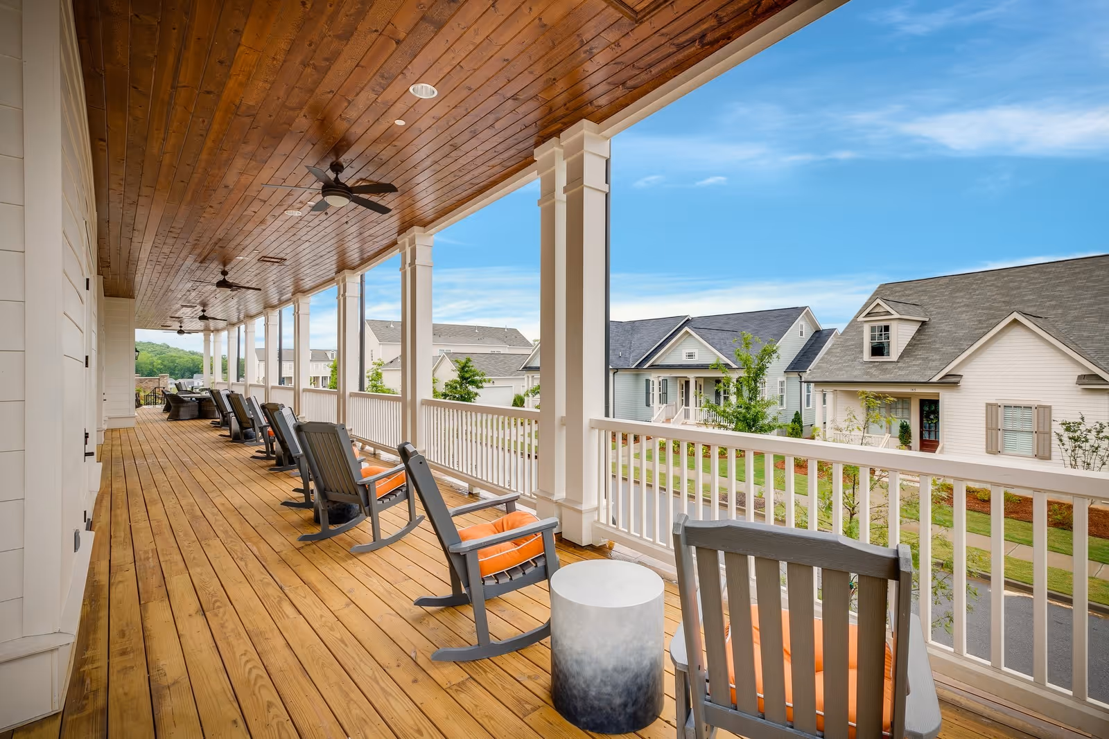 A spacious covered wooden porch with multiple rocking chairs featuring orange cushions, ceiling fans, and a small round table. The porch overlooks a quiet neighborhood with houses and greenery under a blue sky with some clouds.