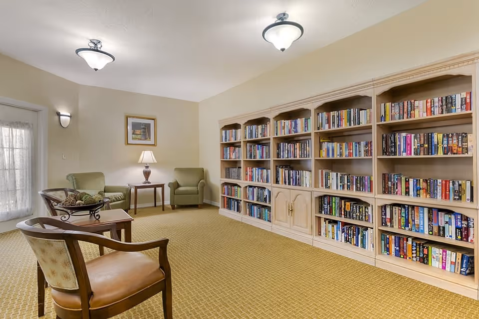 A cozy library room in a senior living facility with a large wooden bookshelf filled with books along one wall. The room has a beige carpet, two green upholstered armchairs, a wooden side table with a lamp, and two wooden chairs with cushioned seats. There is a framed picture on the wall and a glass door with sheer curtains allowing natural light to enter.