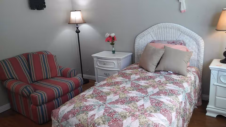 Small bedroom with a single bed covered by a floral quilt, two white nightstands, a floor lamp, and a striped armchair.