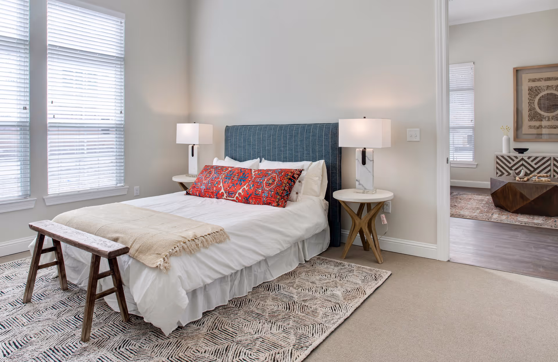 A bright bedroom with a bed featuring a blue upholstered headboard, white bedding, and a long red decorative pillow. There are two white bedside tables with modern lamps on each side of the bed. A wooden bench is placed at the foot of the bed on a patterned rug. Large windows with white blinds allow natural light into the room. An open doorway reveals a glimpse of an adjacent room with a wooden floor, a patterned rug, and a decorative console table with artwork above it.
