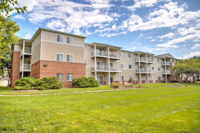 Exterior view of a three-story senior living facility building with beige siding and red brick accents. The building features balconies and is surrounded by a well-maintained green lawn, shrubs, and trees under a partly cloudy blue sky.