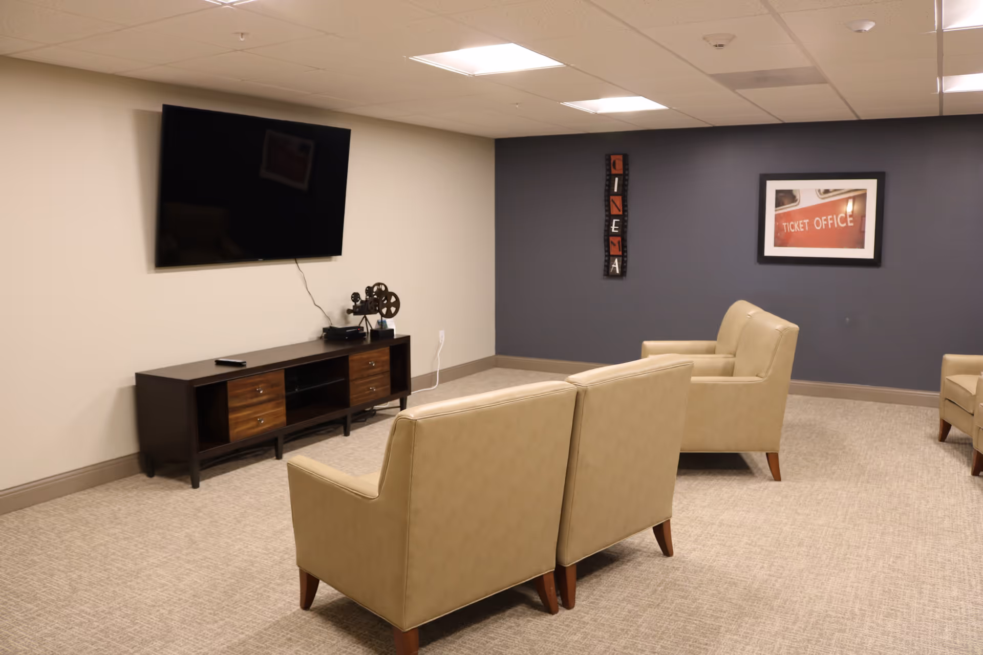 A cozy entertainment room with beige armchairs arranged facing a wall-mounted flat-screen TV. Below the TV is a dark wooden media console with decorative items, including a vintage film projector. The walls are painted beige and dark gray, with a vertical 'CINEMA' sign and a framed picture of a ticket office on the gray wall.
