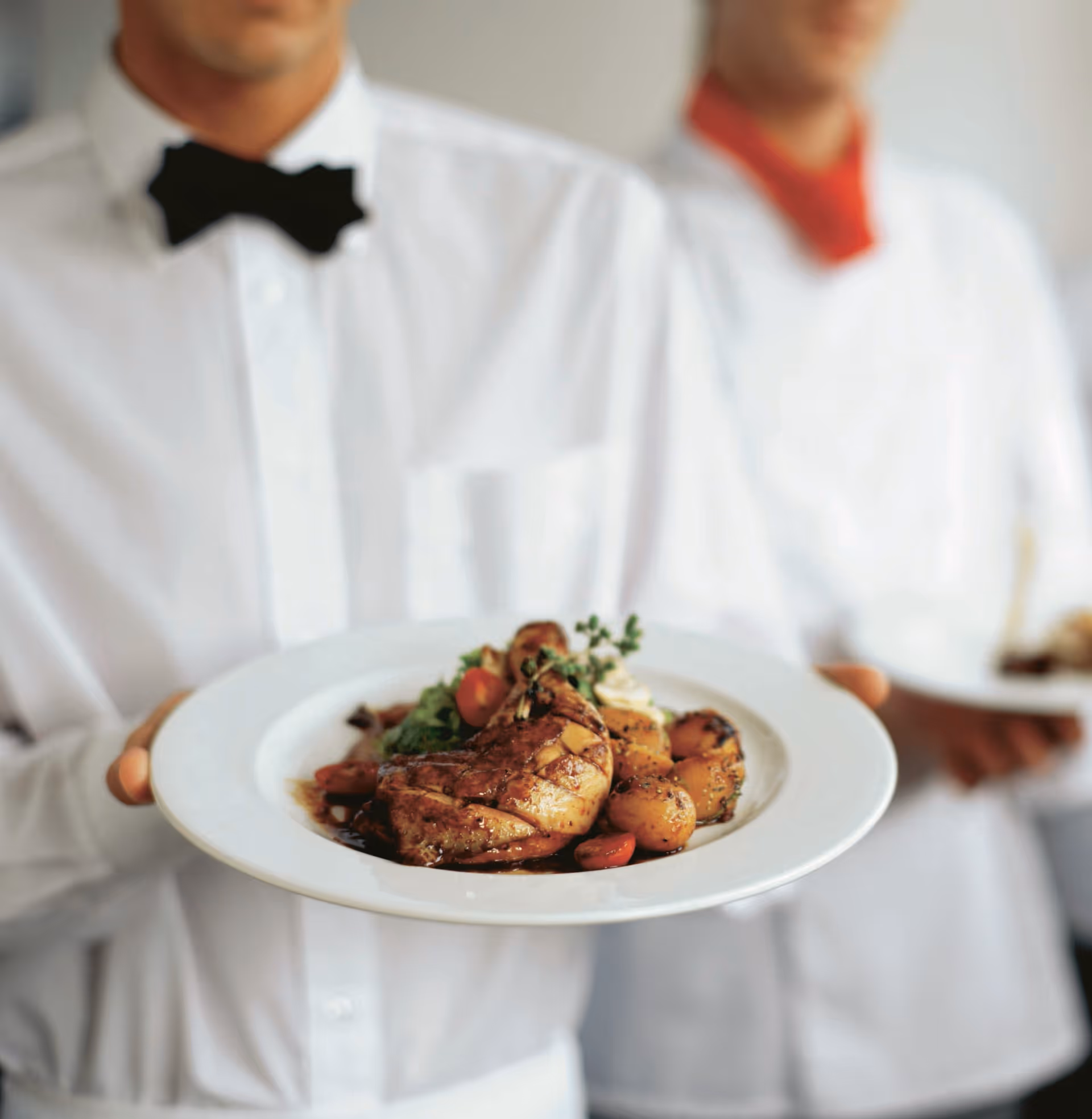 Two waiters dressed in white shirts and bow ties holding plates of food, with the focus on the plate in the foreground featuring a cooked chicken leg, roasted potatoes, cherry tomatoes, and greens.