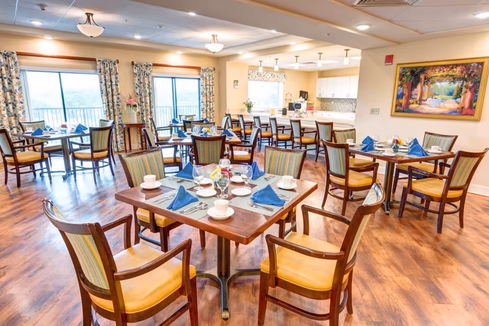 A bright and spacious dining room with multiple wooden tables and chairs arranged neatly. Each table is set with white cups, plates, silverware, and blue folded napkins. Large windows with floral curtains allow natural light to fill the room. In the background, there is a kitchen area with white cabinets and a countertop with bar stools. A colorful painting hangs on the wall to the right.