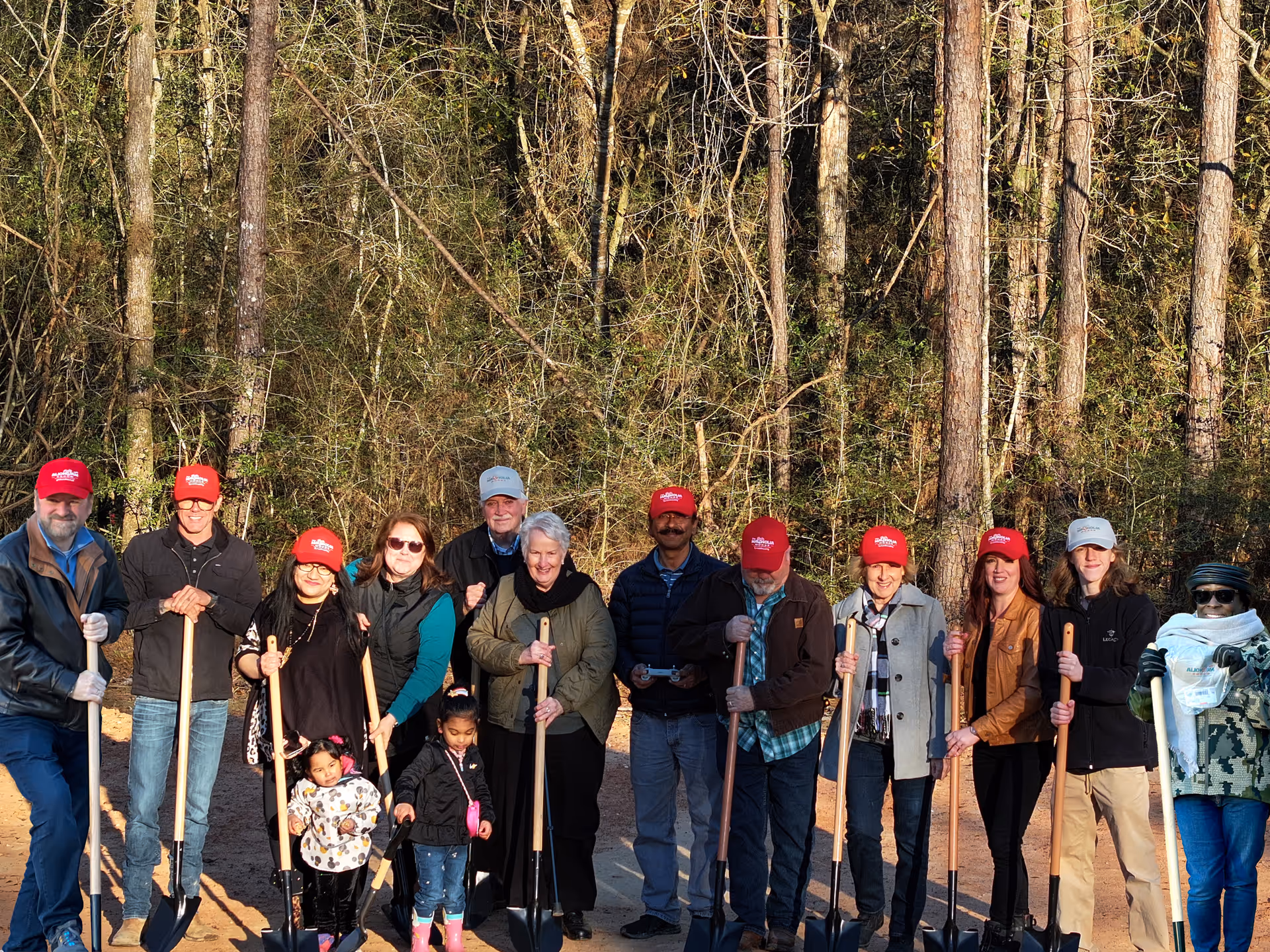 A group of people standing outdoors in front of a wooded area, each holding a shovel. Most are wearing red hats, and there are two young children among the group. The setting appears to be a community or volunteer event.