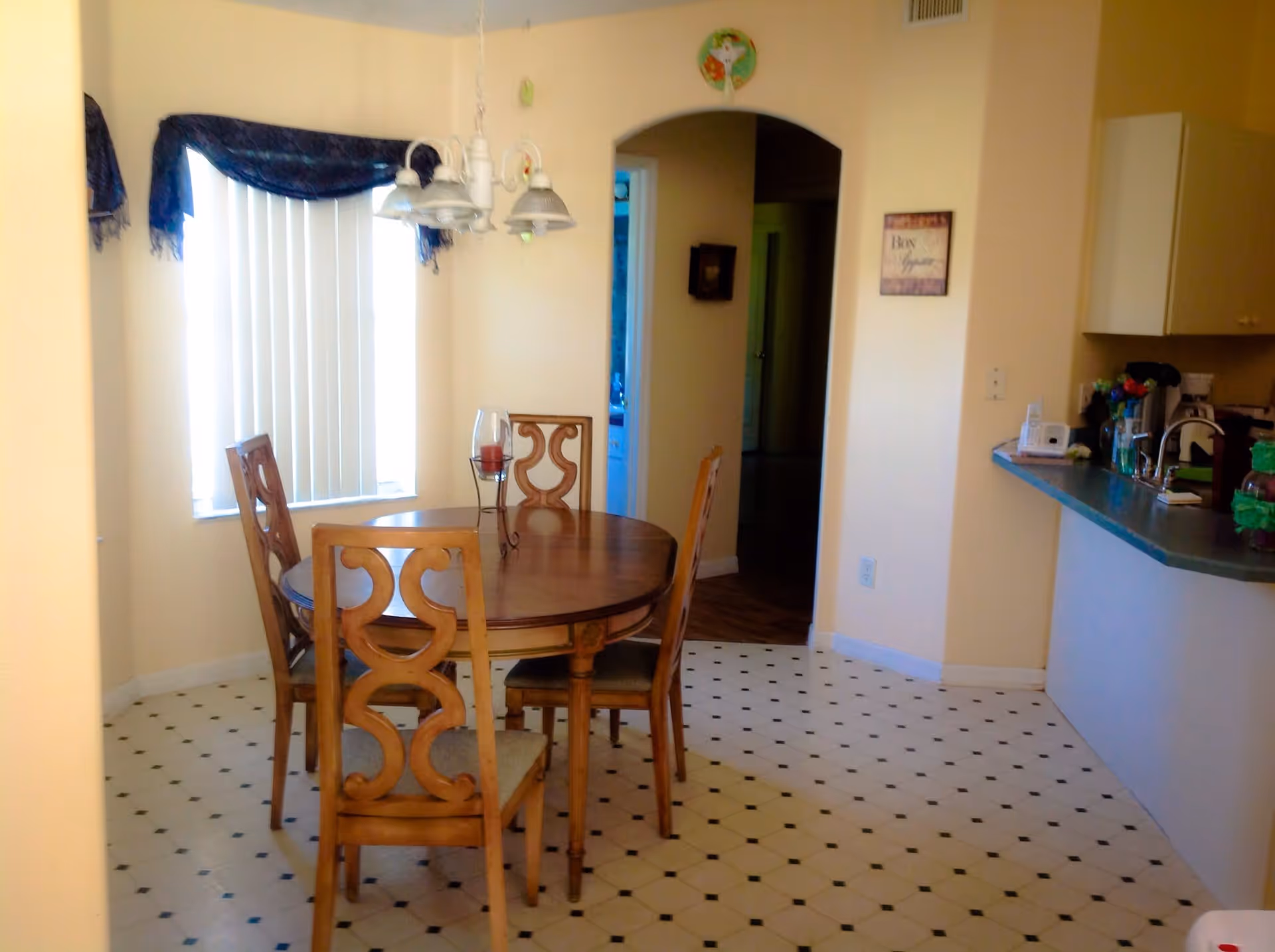 Sunlit dining area with a round wooden table and four chairs beside a pass-through kitchen counter.