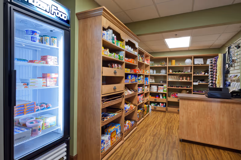 Interior view of a small convenience store or snack shop inside a senior living facility. Wooden shelves are stocked with various snacks, cereals, and other packaged goods. A refrigerated display case on the left contains frozen food items. A wooden counter with a cash register is on the right side. The floor has a wood-like finish and the ceiling has recessed lighting.