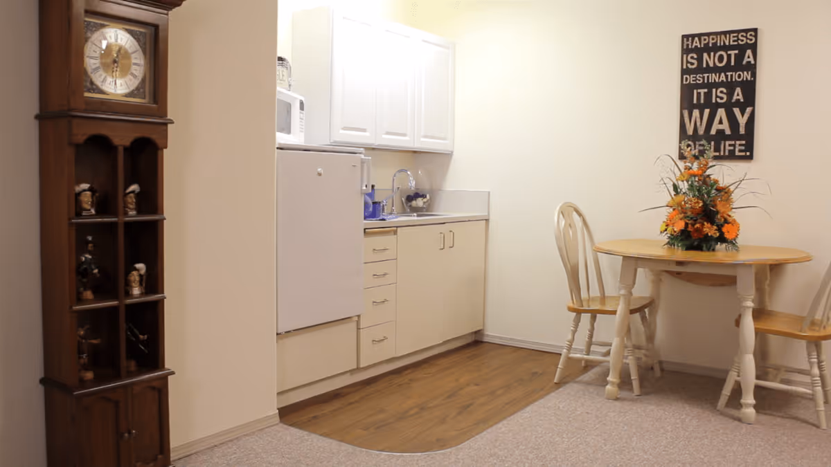 A small kitchen and dining area in an assisted living community. The kitchen has white cabinets, a white refrigerator, a microwave, and a sink. Adjacent to the kitchen is a round wooden dining table with two matching chairs and a floral centerpiece. On the wall above the table is a decorative sign that reads, 'Happiness is not a destination. It is a way of life.' To the left, there is a tall wooden grandfather clock with small decorative figurines on its shelves.