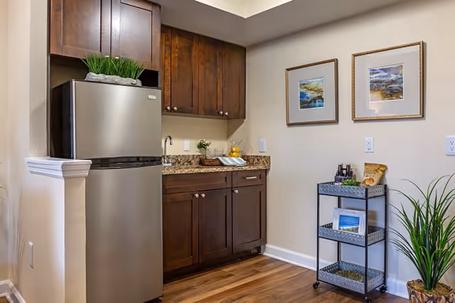 A small kitchen area with dark wooden cabinets, a stainless steel refrigerator, a granite countertop with a sink, and a small metal rolling cart with shelves holding decorative items. Two framed pictures hang on the beige wall, and a potted plant is placed on the floor.