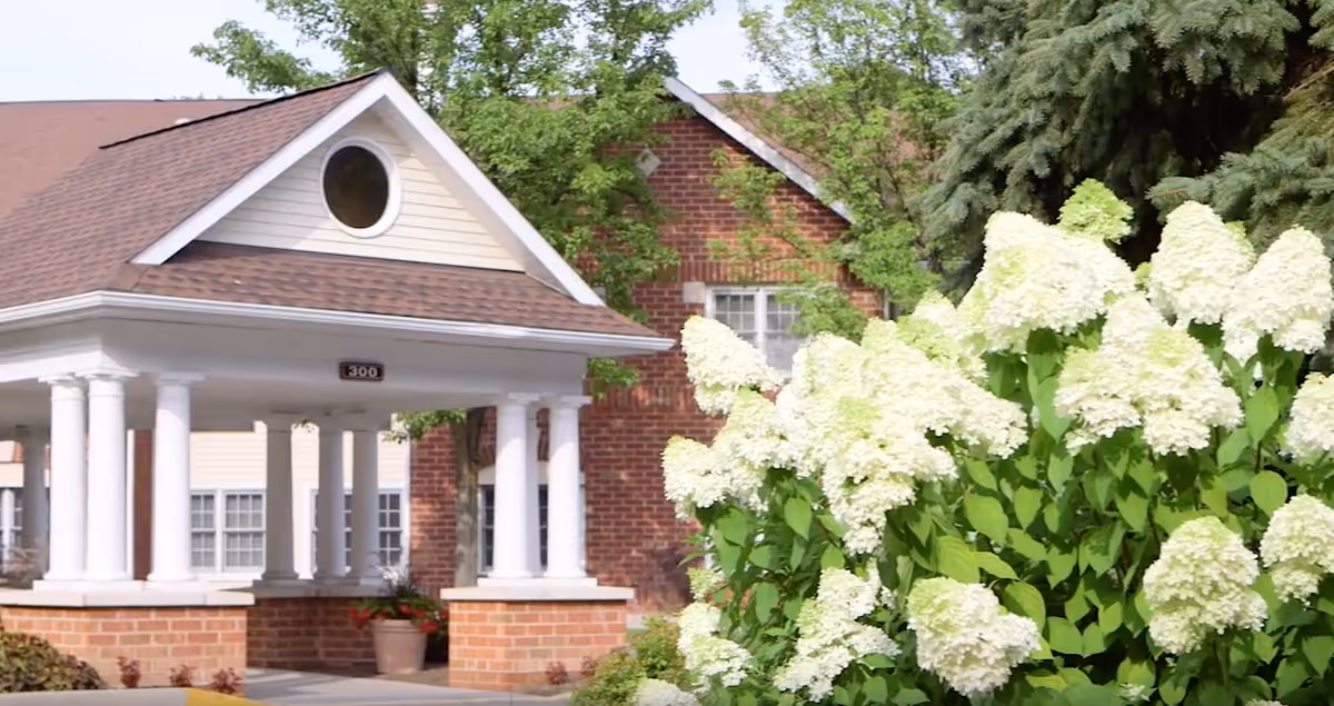 Exterior view of a building entrance with white columns and a small gabled roof, surrounded by green trees and white flowering bushes in the foreground.