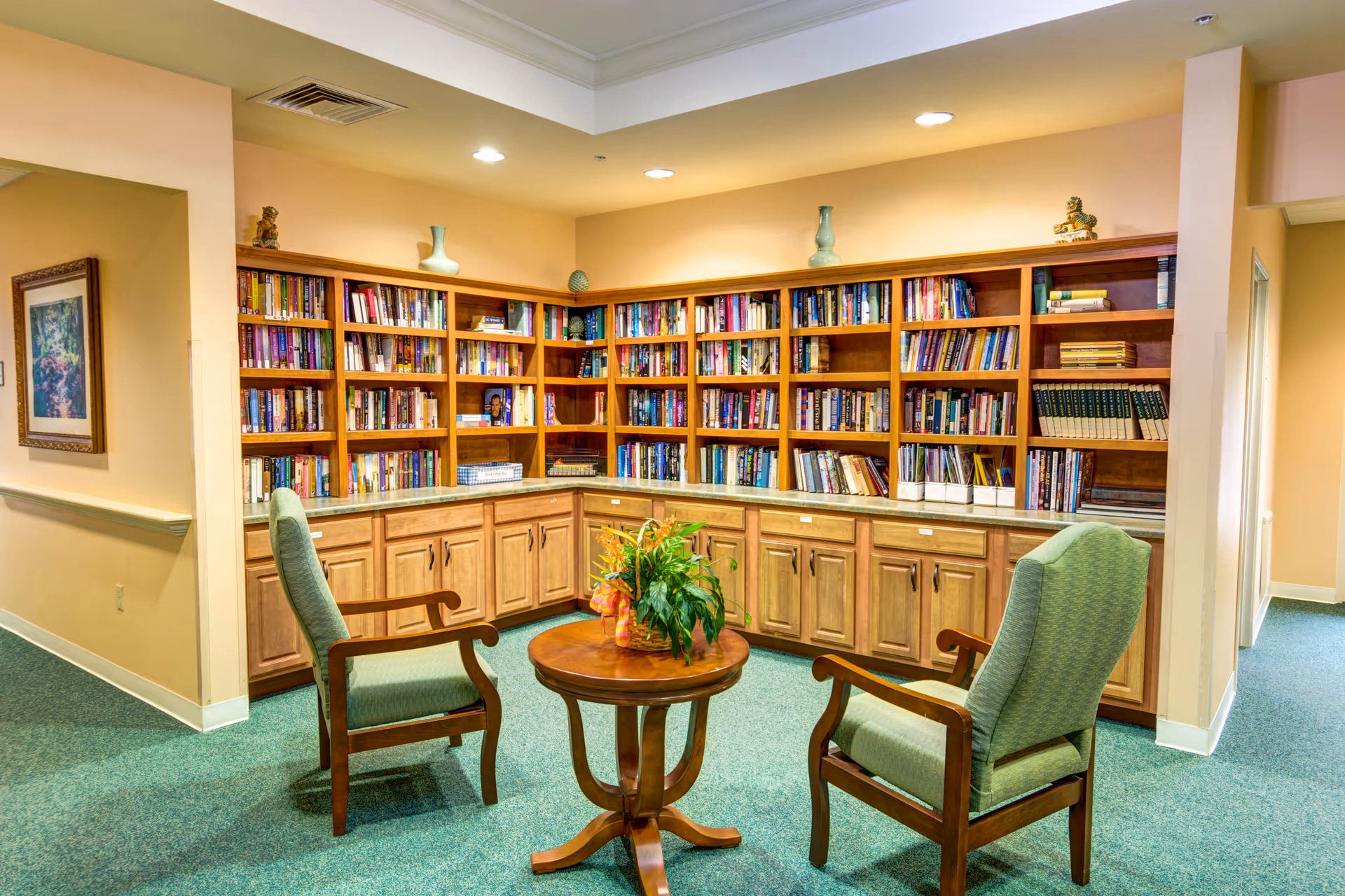 A cozy reading nook in a senior living facility with two green upholstered armchairs facing a round wooden table with a plant centerpiece. Behind the chairs is a large wooden bookshelf filled with books and decorative items. The walls are painted a soft beige, and the floor is carpeted in green.