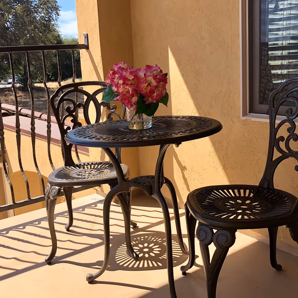 A sunlit balcony with an ornate metal bistro table and two chairs, a vase of pink flowers on the table and a wrought-iron railing.
