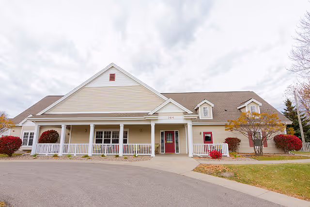 Exterior view of a single-story assisted living facility building with beige siding, a brown roof, white railings on the front porch, and red doors and bushes. The sky is cloudy and there is a paved driveway in front.