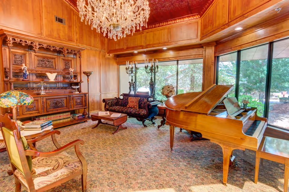 Ornately furnished common room with a grand piano, carved wooden cabinetry, chandelier, seating, and large windows.