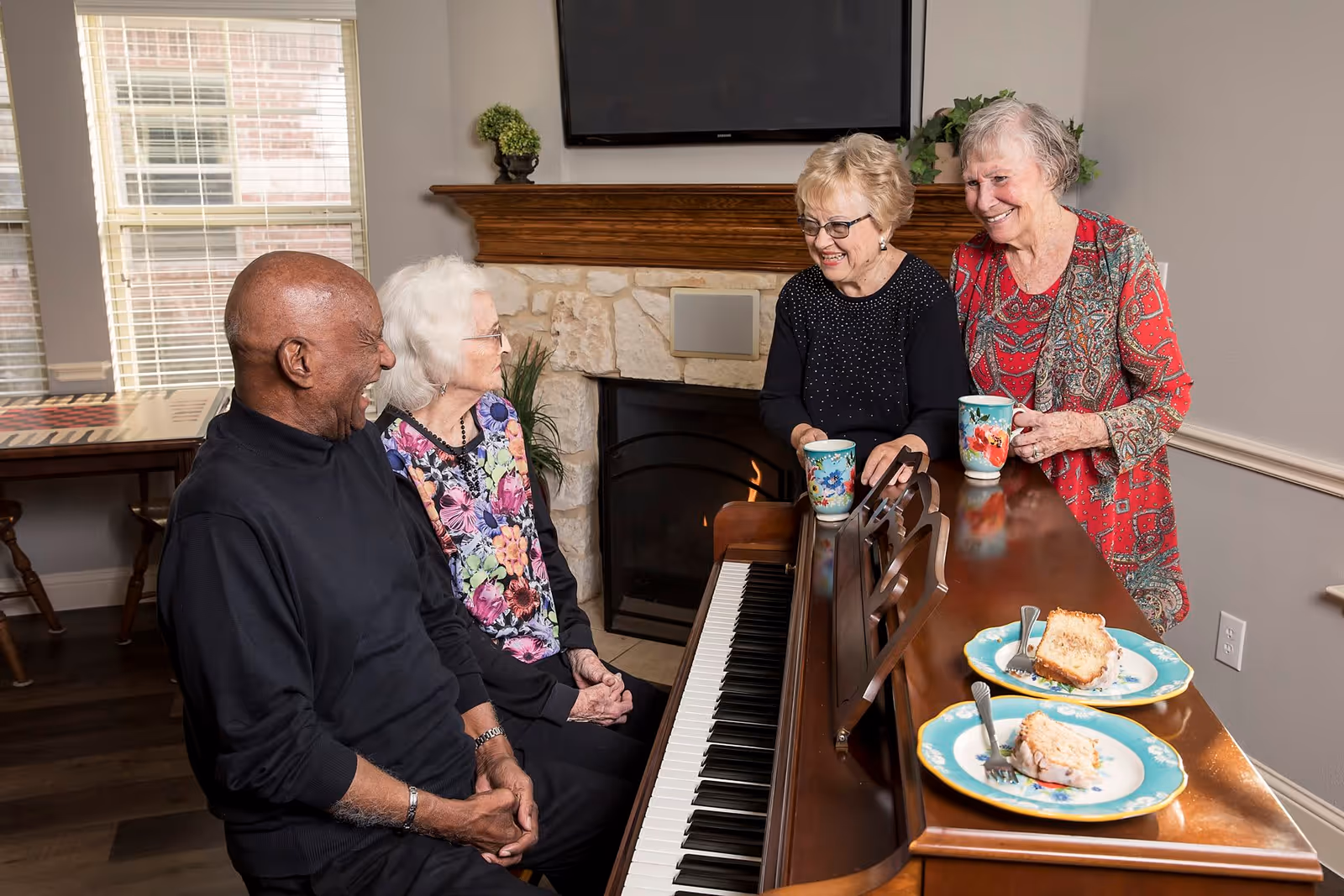 Four elderly people gathered around a piano in a cozy living room. Two women stand on the right side of the piano holding colorful mugs, while a man and a woman sit on the left side, smiling and engaging in conversation. There are two plates with slices of cake and forks on the piano. The room features a stone fireplace, a mounted TV above it, and a window with blinds letting in natural light.