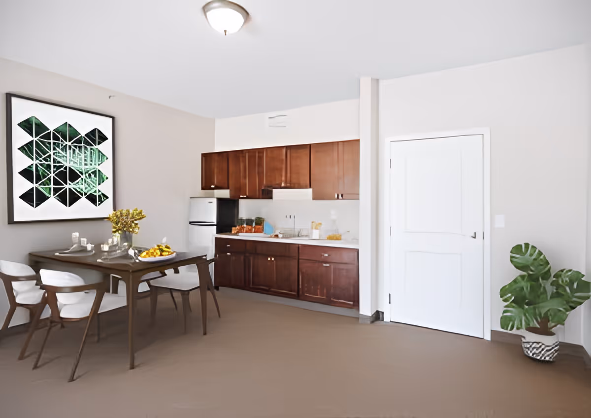 Dining area with a wooden table and chairs in front of a kitchenette with dark cabinets, a small refrigerator, wall art, and a potted plant by a closed door.