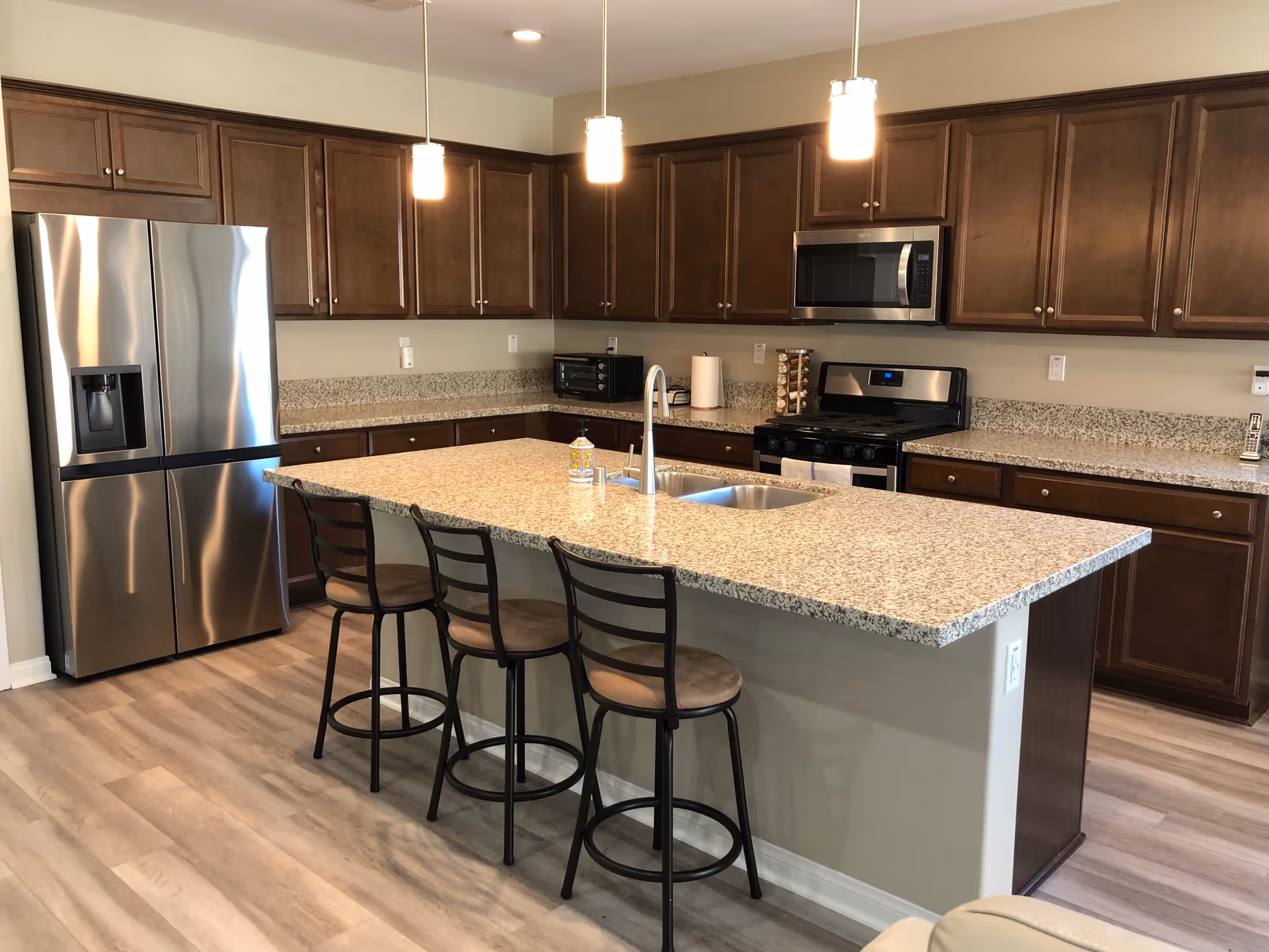Modern kitchen with granite countertops, a large island with three bar stools, stainless steel refrigerator, stove, microwave, and wooden cabinets.