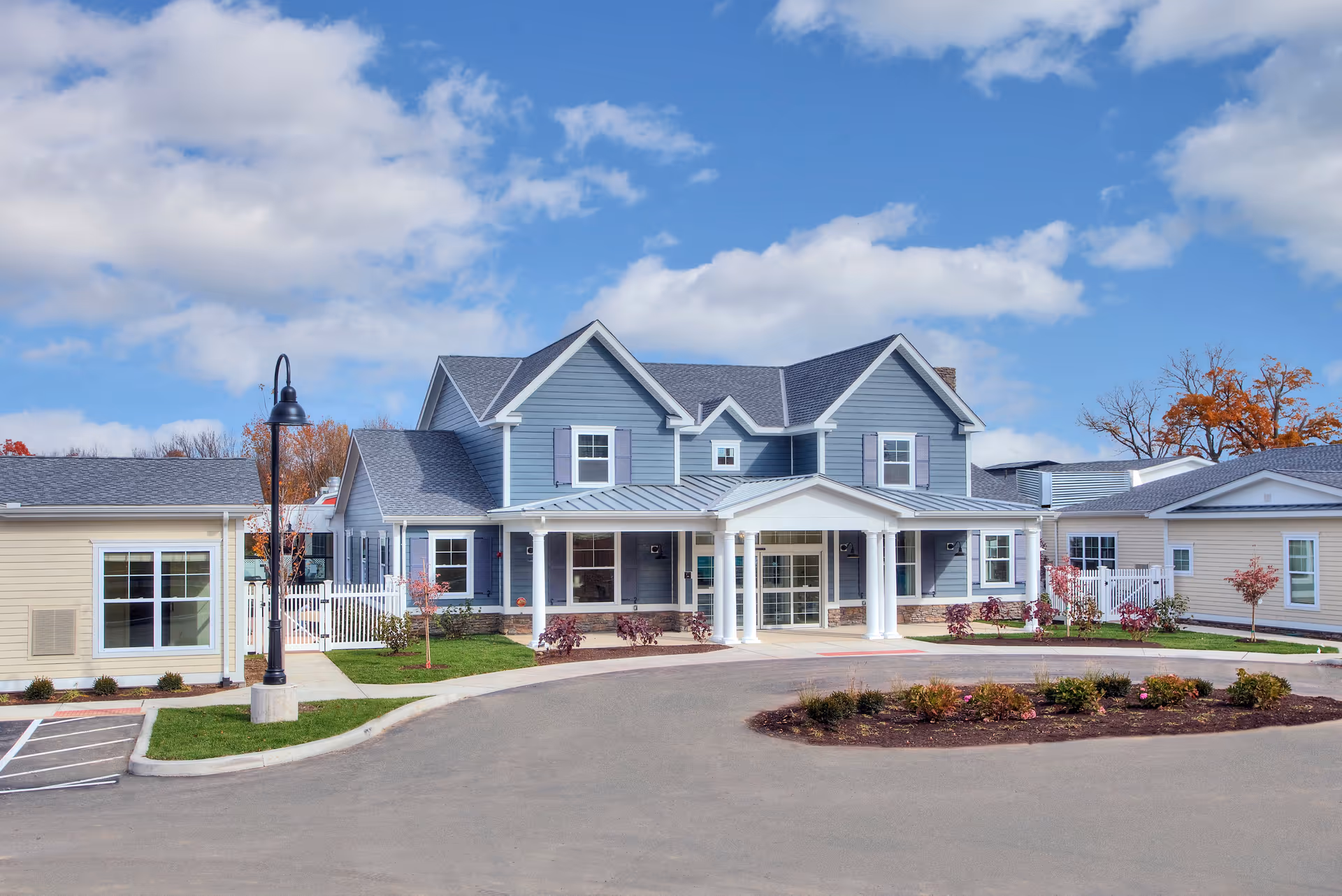 Front exterior view of a senior living facility named Church Hill Village featuring a large blue and white building with a covered entrance supported by white columns, surrounded by a circular driveway and landscaped with small trees and shrubs under a partly cloudy sky.