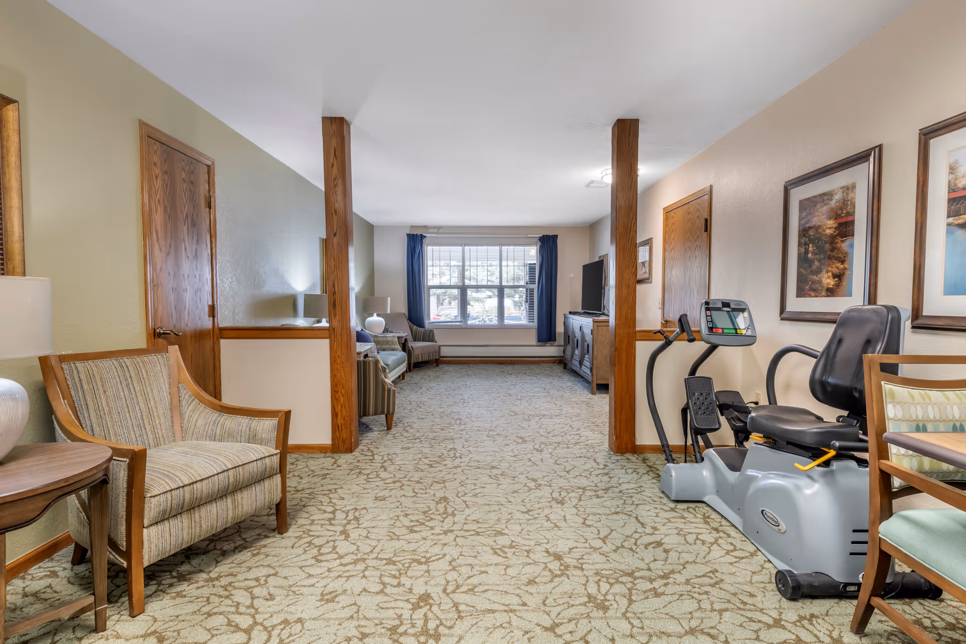 A long room with patterned carpet and beige walls featuring wooden trim. On the left side, there are two armchairs and a small table with a lamp. On the right side, there is a recumbent exercise bike next to a wooden chair with a cushion. At the far end of the room, there is a large window with blue curtains, a TV on a wooden cabinet, and additional seating including a sofa and armchair.