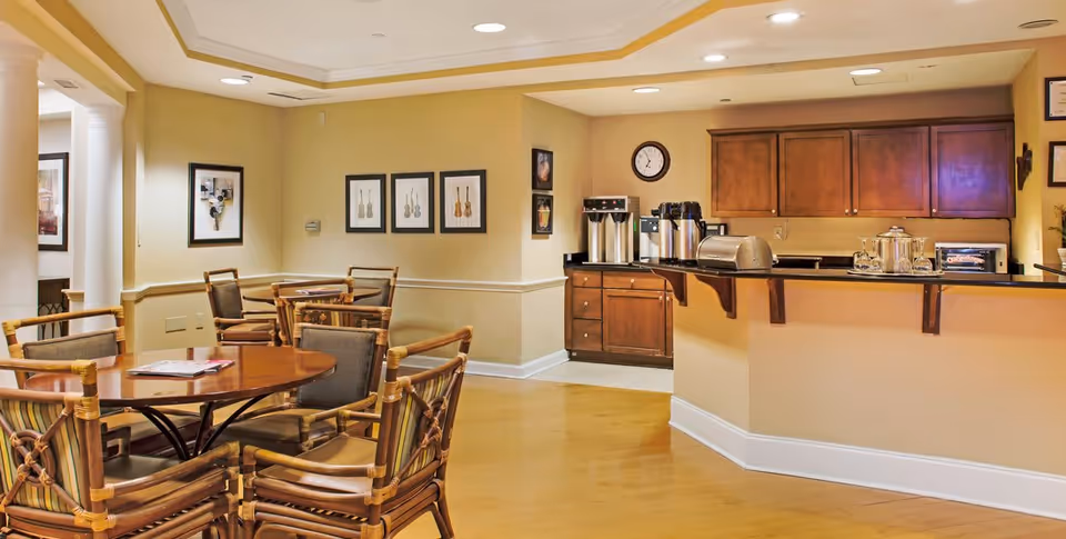 A cozy senior living common area with wooden tables and chairs arranged for seating. The room features a small kitchenette with wooden cabinets, a counter with coffee dispensers, a toaster, and a microwave. The walls are decorated with framed artwork and a clock, and the floor is light wood.