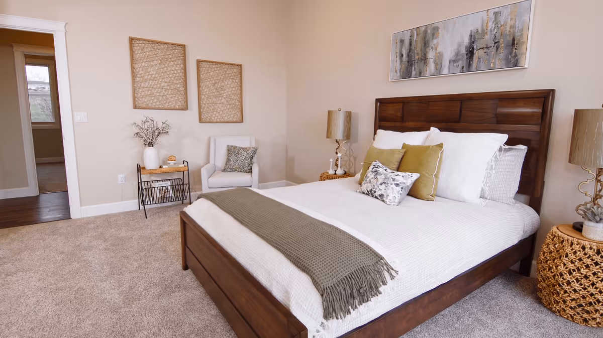 A cozy bedroom with a wooden bed frame, white bedding, and decorative pillows. There is a green throw blanket at the foot of the bed. On the right side, there is a woven nightstand with a lamp. On the left side, there is a small table with a vase and a white armchair with a patterned cushion. Two woven wall hangings and a large abstract painting decorate the walls. The room has beige walls and carpeted flooring.