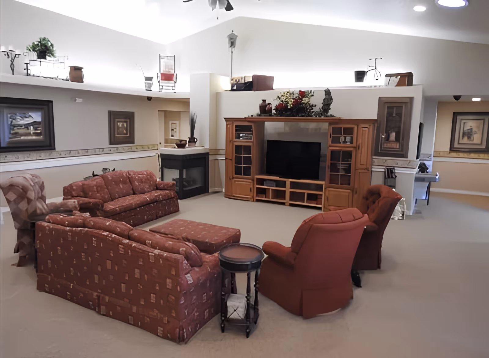 Open communal living room with patterned sofas, red armchairs, ottoman, a wooden entertainment center and fireplace.