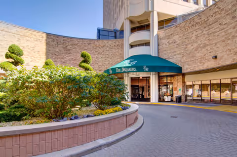 Entrance to The Breakers at Edgewater Beach with a green awning displaying the facility's name, surrounded by a curved brick wall and landscaped greenery including bushes and topiary trees.