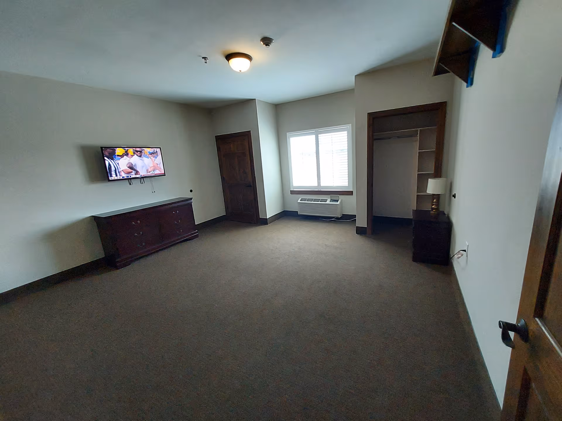 Carpeted senior living bedroom with a wall-mounted TV above a dresser, an open closet, window air conditioner, and a nightstand.