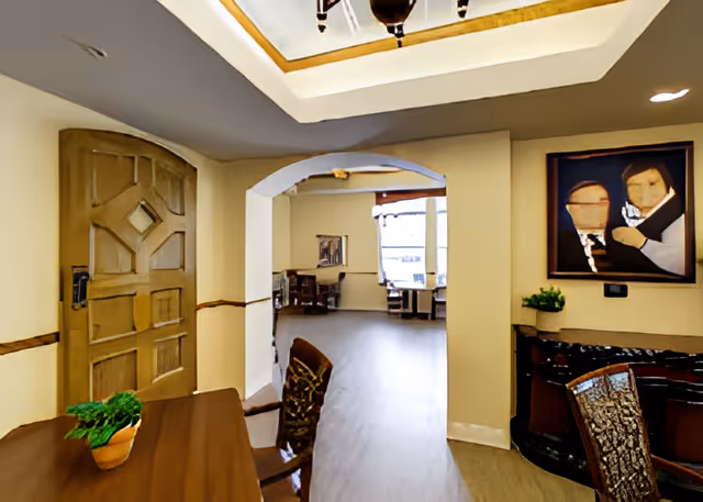 Interior view of a senior living facility showing a wooden door on the left, a table with a small potted plant, and chairs. The room has beige walls with a decorative wooden trim and a ceiling with recessed lighting. An archway leads to another room with more tables and chairs near large windows. A framed painting of two abstract human figures hangs on the right wall above a dark wooden cabinet with another small plant.