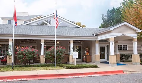 Front entrance of a senior living building with a covered portico, two flagpoles, landscaping, and the address '6015' above the doorway.