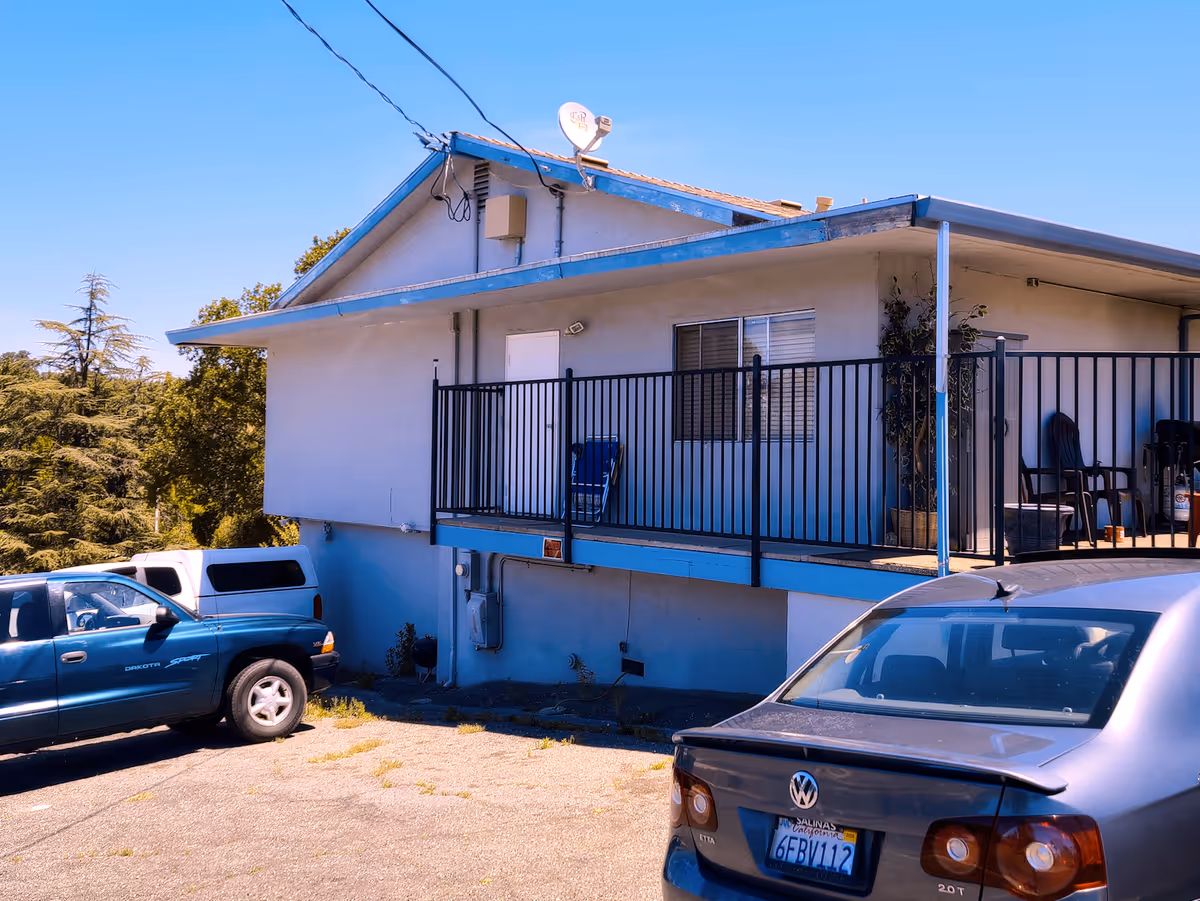Exterior view of a single-story building with a blue roof and railing, a small balcony with chairs, and two parked cars in front under a clear blue sky.