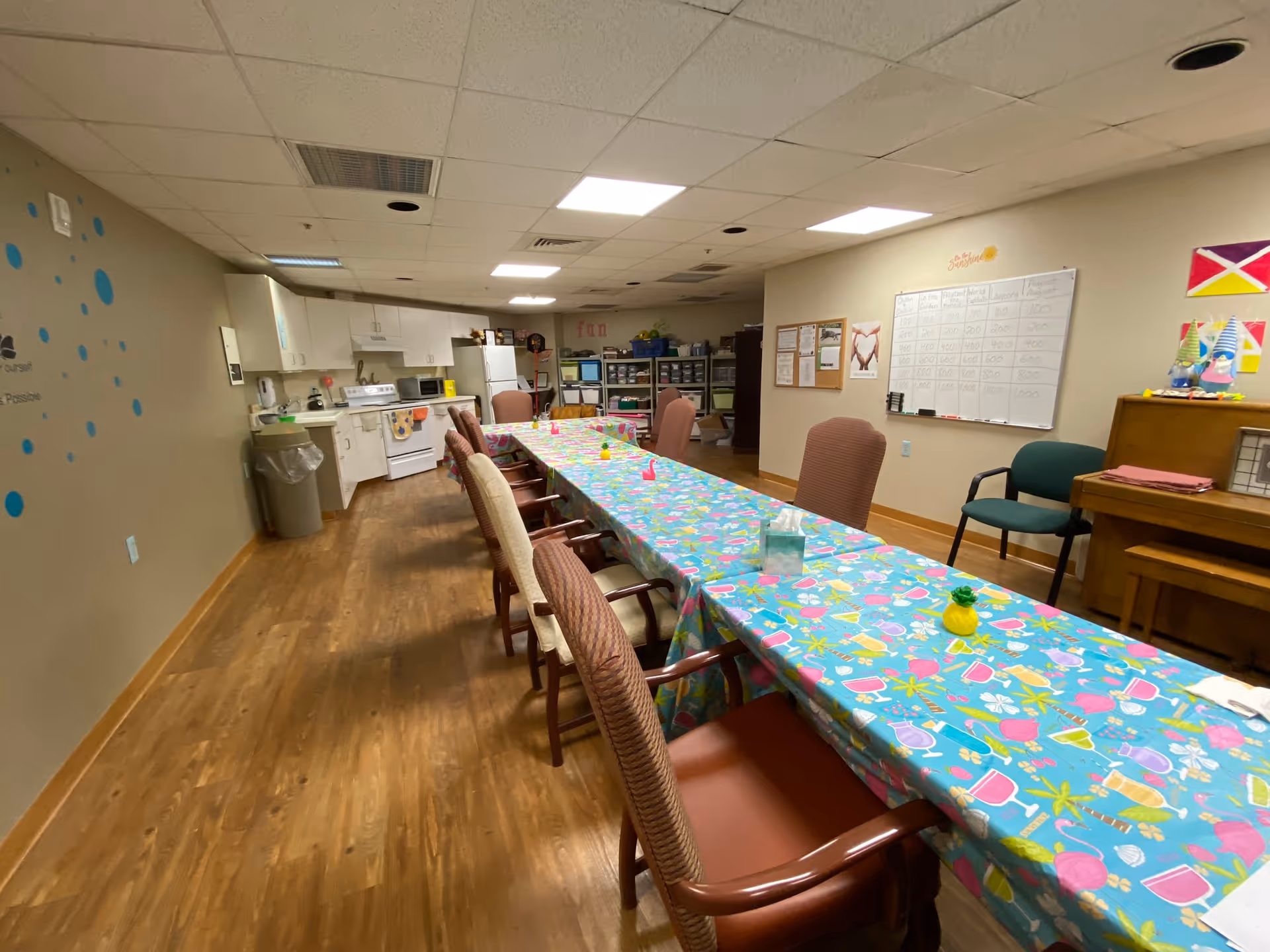 A long table covered with a colorful tablecloth featuring tropical drinks and palm trees, surrounded by various chairs in a room with wood flooring. In the background, there is a kitchen area with white cabinets, a stove, microwave, and refrigerator. On the right wall, there is a whiteboard with a chart and some decorations, including a piano and chairs.