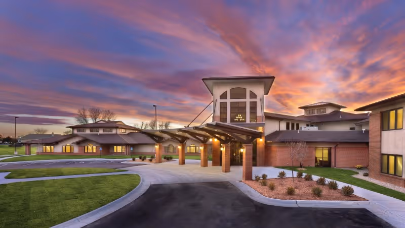 Front entrance of an assisted living facility with a covered porte-cochere, lit windows, and a dramatic sunset sky.