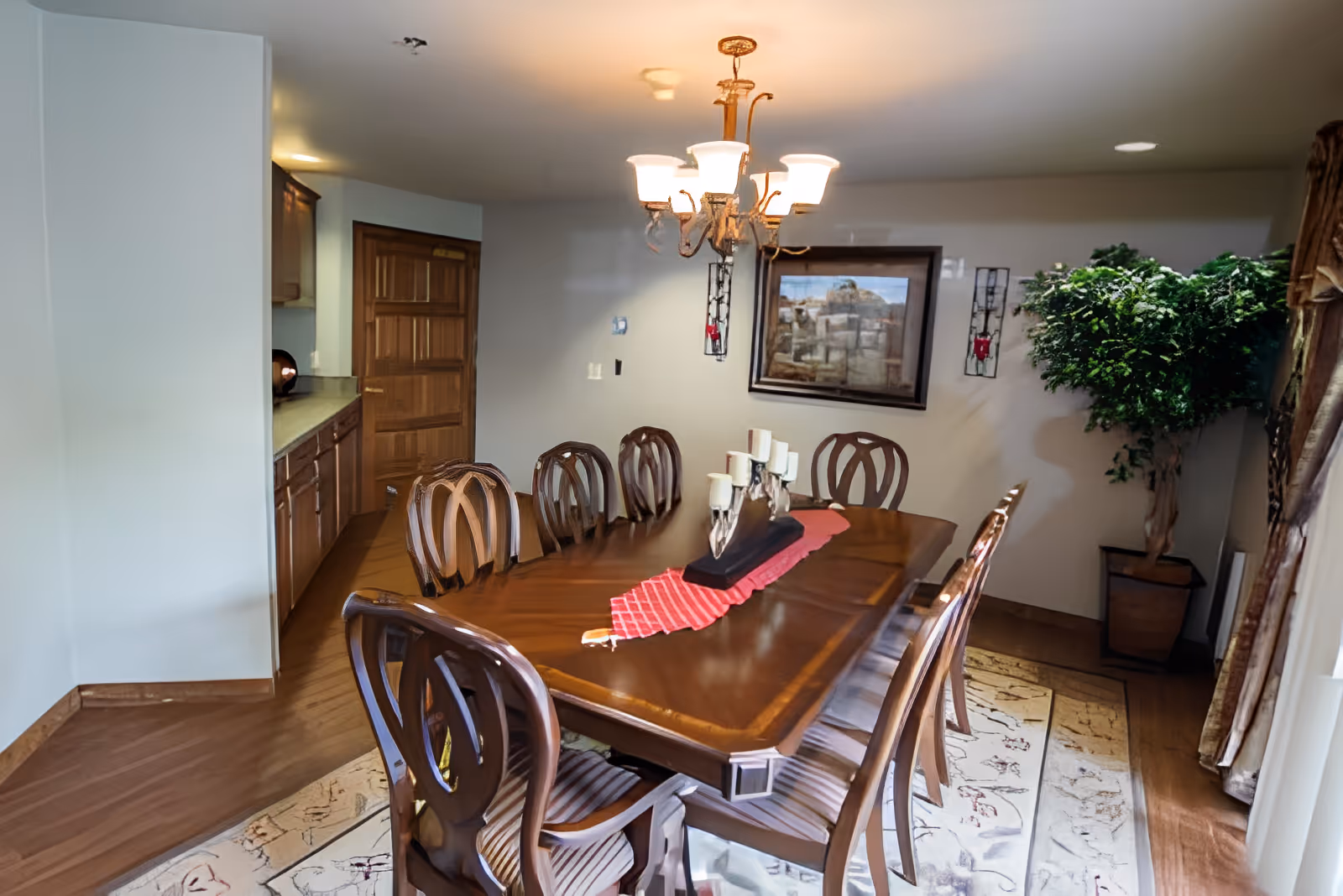 Formal dining room with a long wooden table and chairs under a chandelier, a side cabinet, framed artwork, and a potted plant.