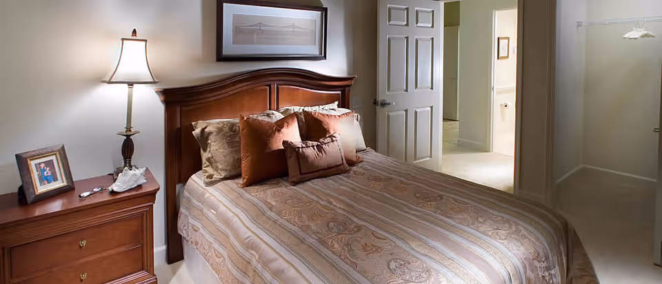 Bedroom with a made bed and wooden headboard, a nightstand with a lamp and framed photo, and open doors leading to a bathroom and closet.