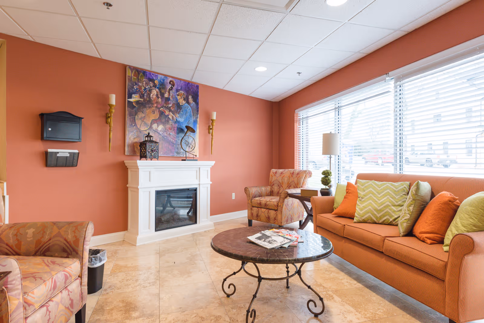 A cozy living room area with a large window covered by blinds letting in natural light. The room features an orange sofa with multiple colorful cushions, two patterned armchairs, a round coffee table with magazines on it, a white fireplace with decorative items on the mantel, and a colorful painting above the fireplace. The walls are painted a warm terracotta color and the floor is tiled.