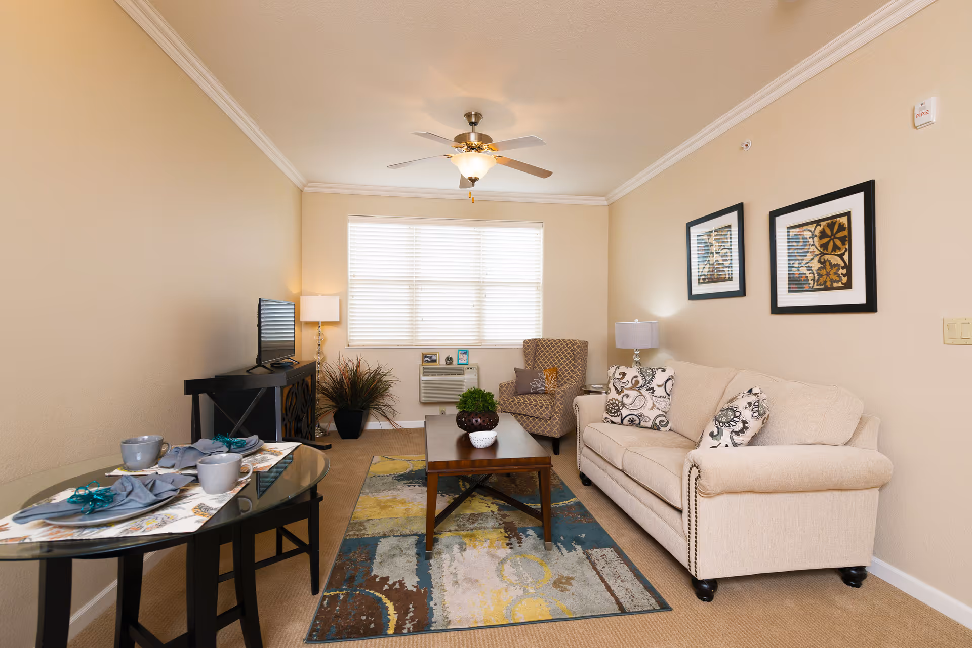 A cozy living room with a beige sofa adorned with patterned cushions, a patterned armchair, a wooden coffee table on a multicolored rug, a TV on a black stand, and a small glass dining table set with two cups and plates. The room has beige walls, a ceiling fan with light, and a window with white blinds letting in natural light.