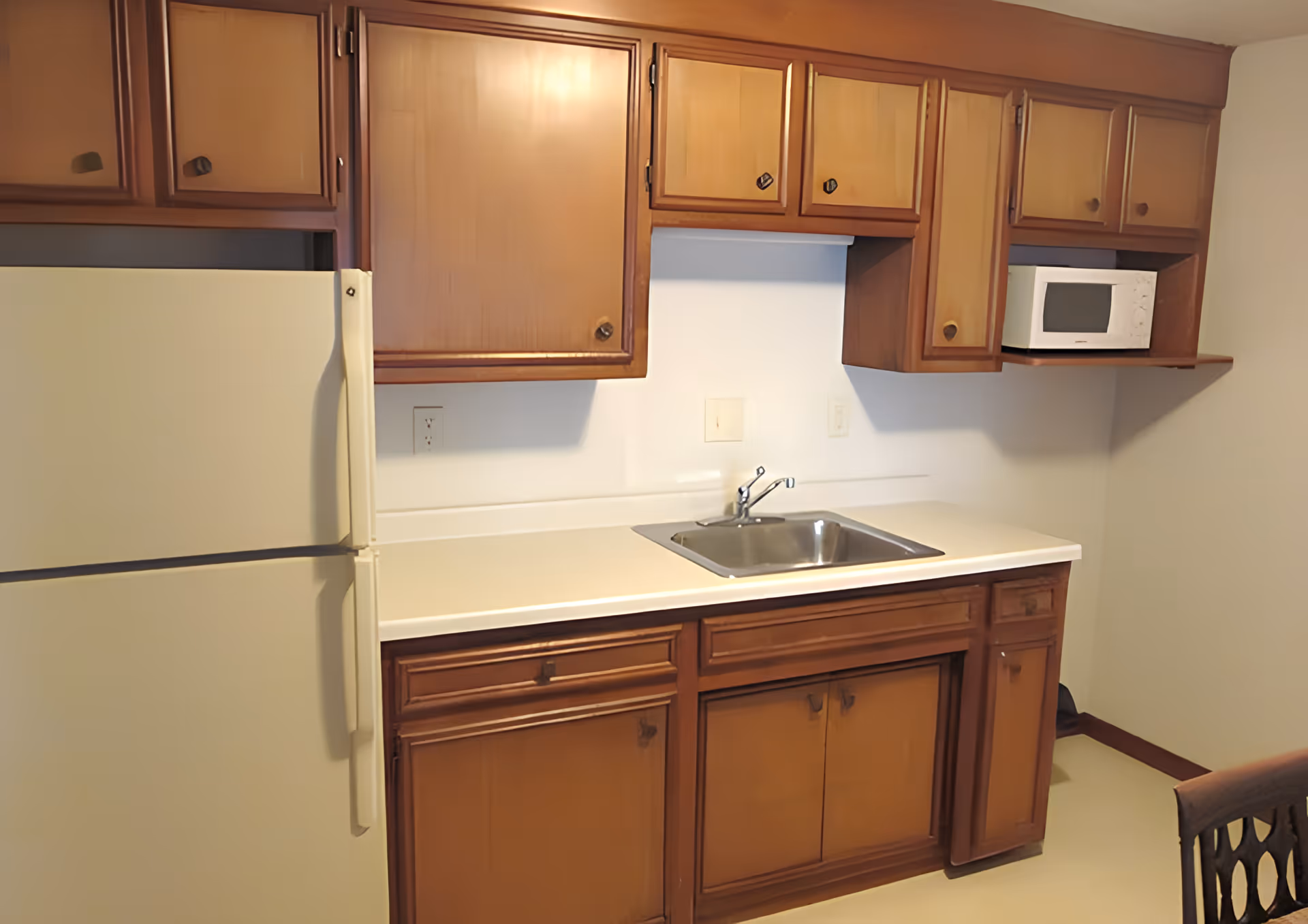 A small kitchen area with wooden cabinets, a white refrigerator, a stainless steel sink, a white countertop, and a white microwave placed on a wooden shelf.