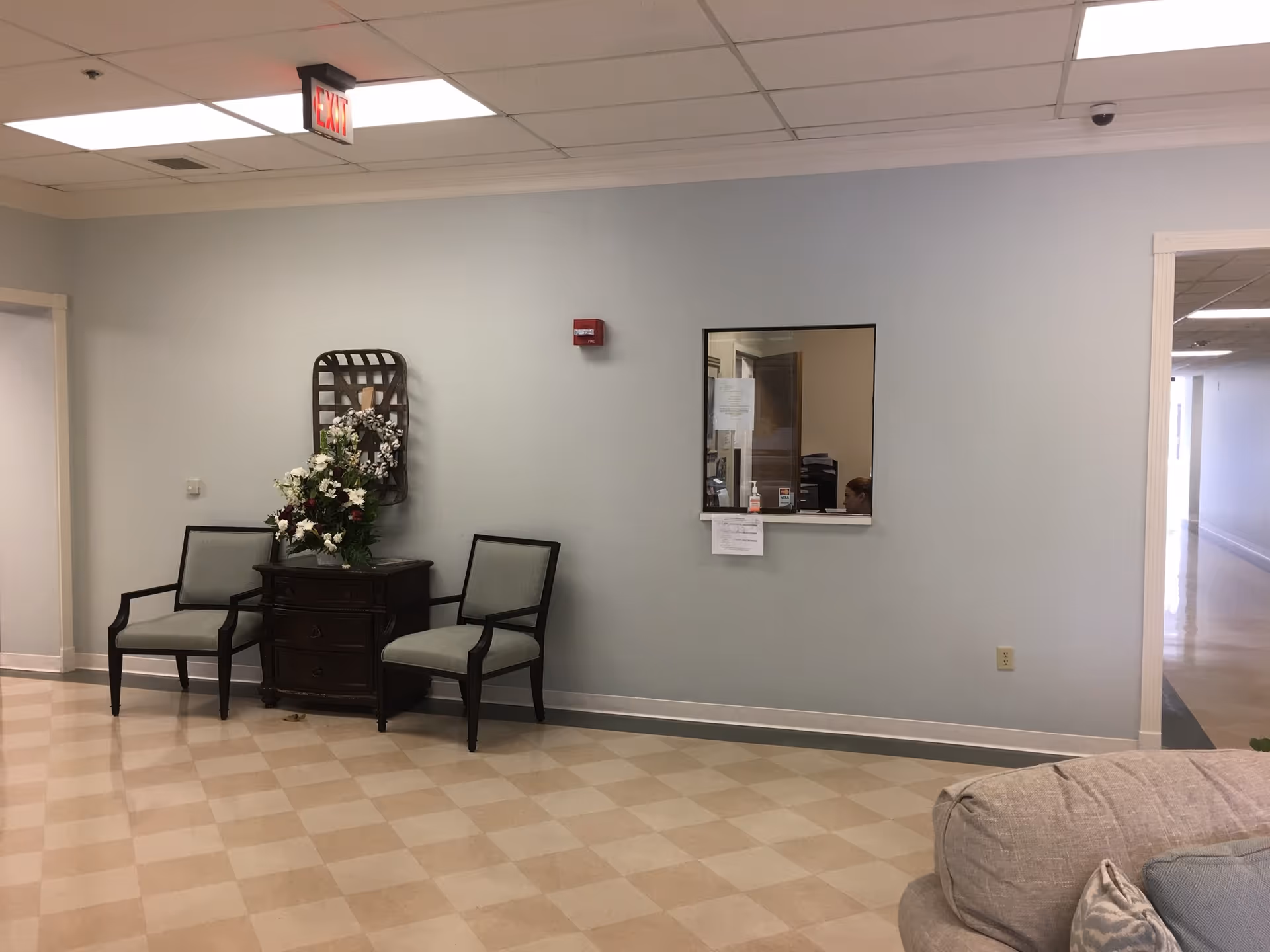 A waiting area inside Westside Terrace Healthcare with two gray cushioned chairs flanking a small dark wooden table topped with a floral arrangement. There is a windowed reception area on the wall and an exit sign on the ceiling. The floor has a checkered pattern and a beige couch is partially visible in the foreground.