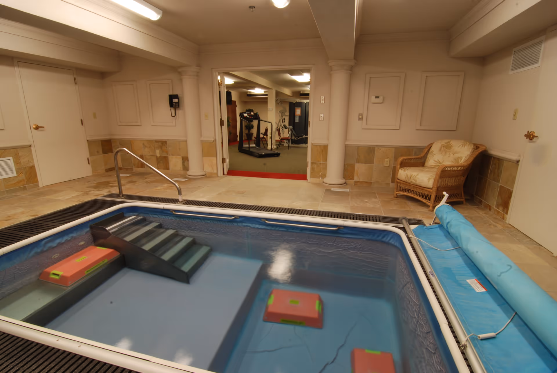 Indoor therapy pool with steps and floating equipment in a tiled room. A wicker chair with a cushion is placed against the wall. Through an open doorway, a fitness room with exercise equipment including a treadmill and weight machine is visible.