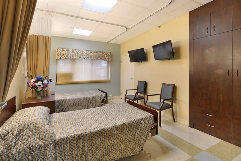 A senior living facility bedroom with two single beds covered in patterned bedspreads. There is a window with a decorative valance, a nightstand with a vase of flowers, two chairs, two wall-mounted flat-screen TVs, and a large wooden wardrobe.