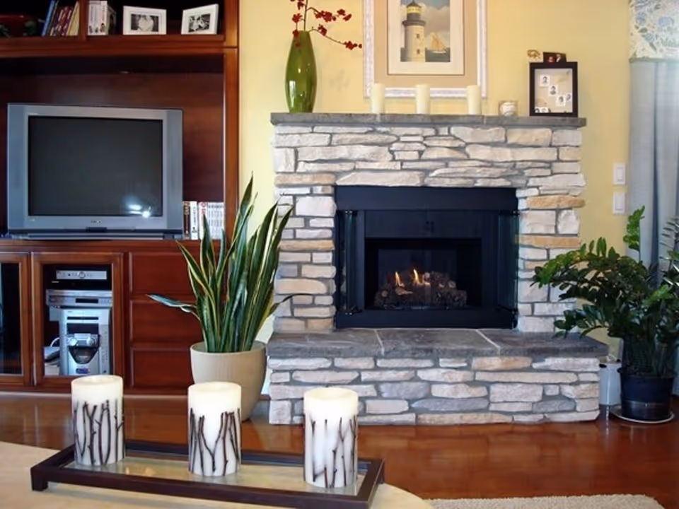 A cozy living room featuring a stone fireplace with a small fire burning inside. On the mantel above the fireplace are three candles, a green vase with red flowers, and a framed picture of a lighthouse. To the left of the fireplace is a wooden entertainment center with a TV, stereo system, and some books. There are two potted plants placed near the fireplace and entertainment center. In the foreground, there is a glass coffee table with three decorative candles on a tray.