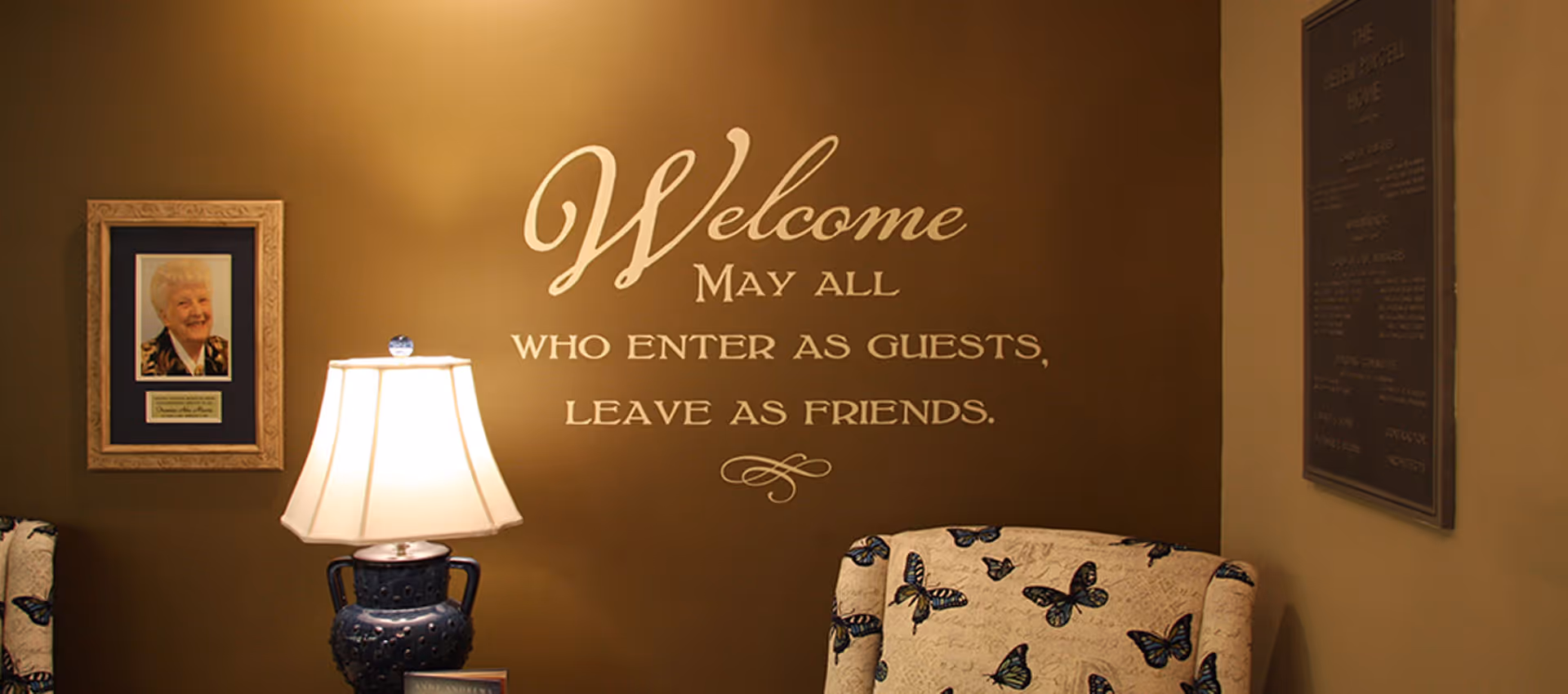 Cozy reception seating area with a lamp, framed portrait, butterfly-upholstered chair, and a wall decal reading 'Welcome — May all who enter as guests, leave as friends.'