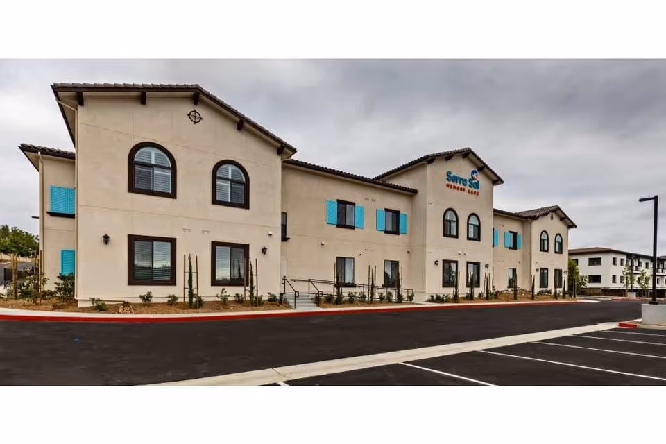 Exterior view of Serra Sol Memory Care facility showing a two-story beige building with blue window shutters, arched windows, and a tiled roof under a cloudy sky. The parking lot is visible in the foreground.