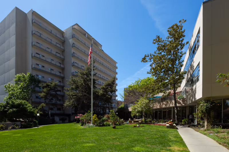 Outdoor view of Piedmont Gardens facility showing a tall multi-story building on the left and a lower building on the right, with a well-maintained green lawn, trees, and an American flag on a flagpole in the center under a clear blue sky.