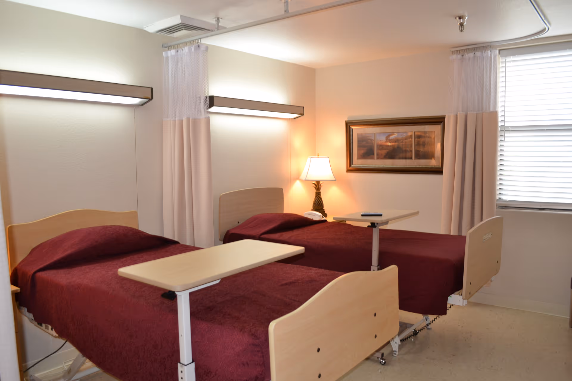 A nursing center room with two hospital-style beds covered in dark red blankets. Each bed has an adjustable overbed table. There is a lamp on a small table between the beds, a framed landscape picture on the wall, and a window with blinds and a privacy curtain.