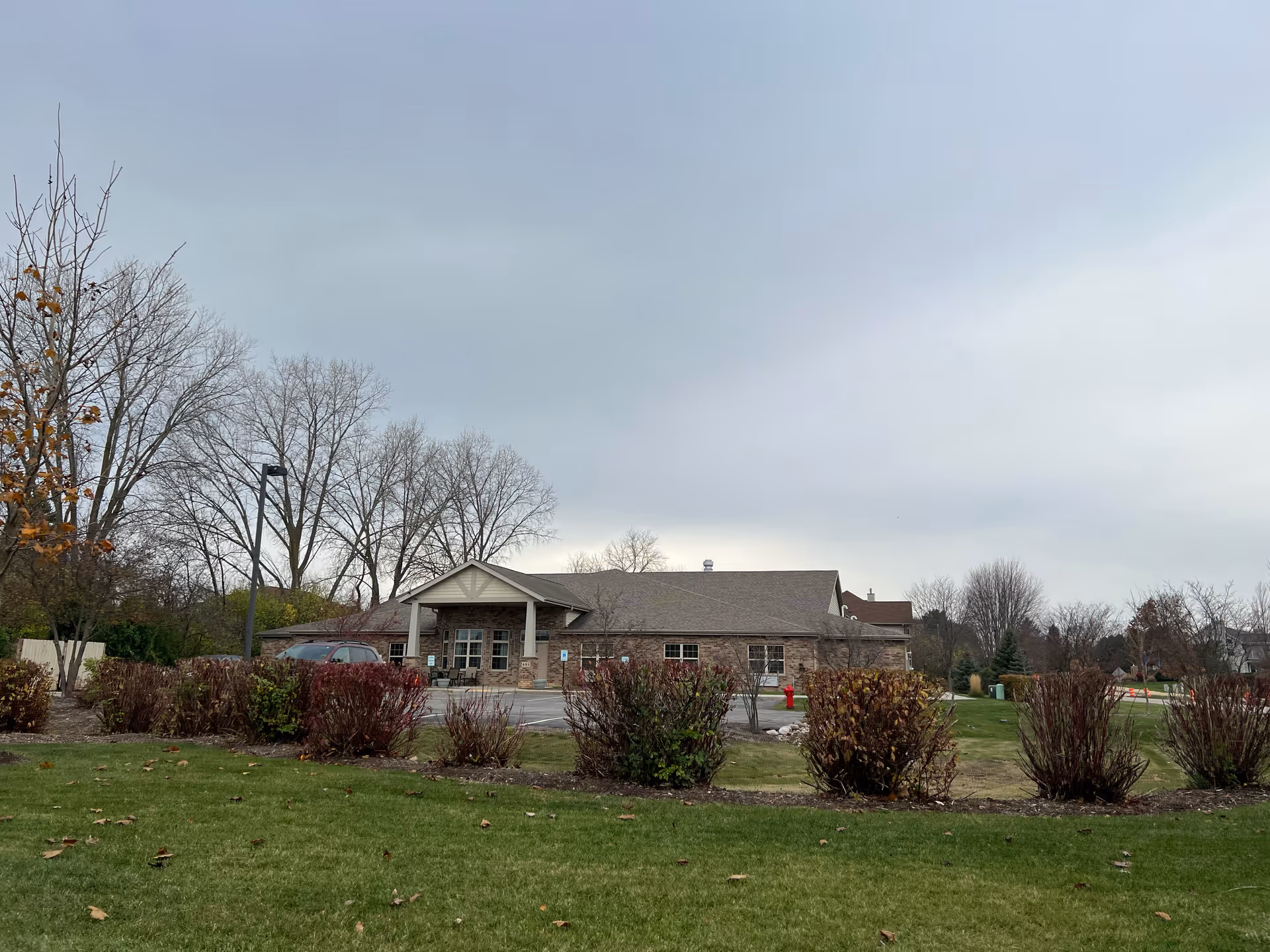 Exterior view of a single-story brick building with a covered entrance and several windows, surrounded by a lawn, bushes, and leafless trees under a cloudy sky.