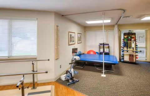 Rehabilitation therapy room with a blue treatment table, exercise equipment, and windows.