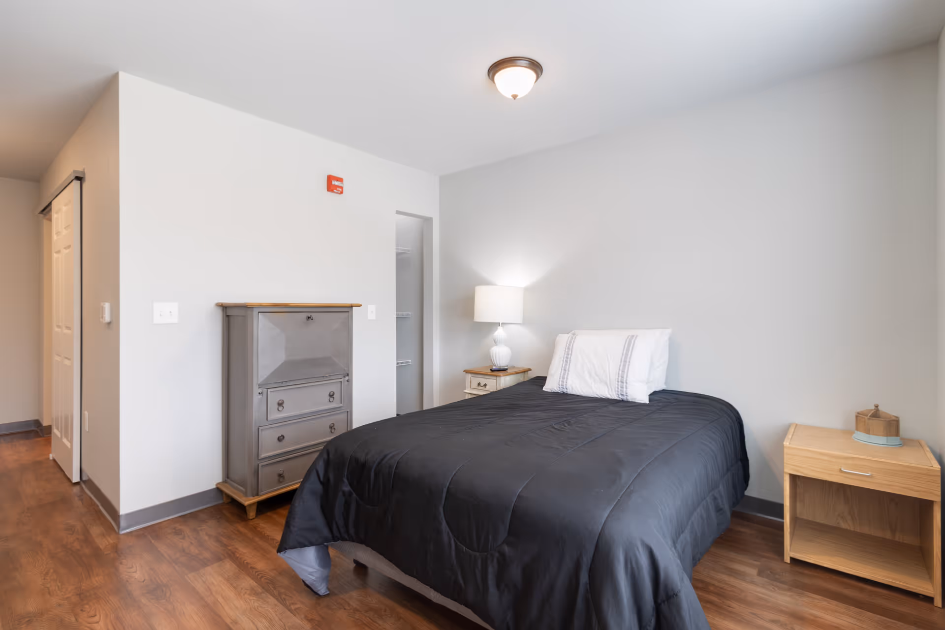 A simple bedroom with a bed covered in a black comforter and a white pillow. There is a wooden nightstand with a lamp on the right side of the bed and a small wooden cabinet with drawers on the left side. The room has light gray walls, a wooden floor, and a ceiling light fixture. A closet with sliding doors is visible in the background.