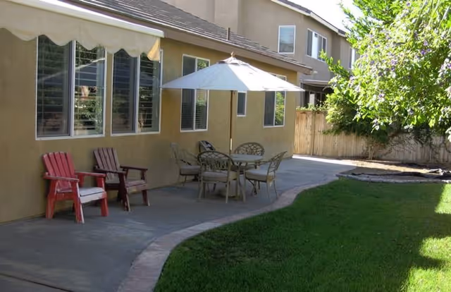 Backyard patio with a round table and umbrella, red chairs, and a grassy lawn beside a beige two-story building.